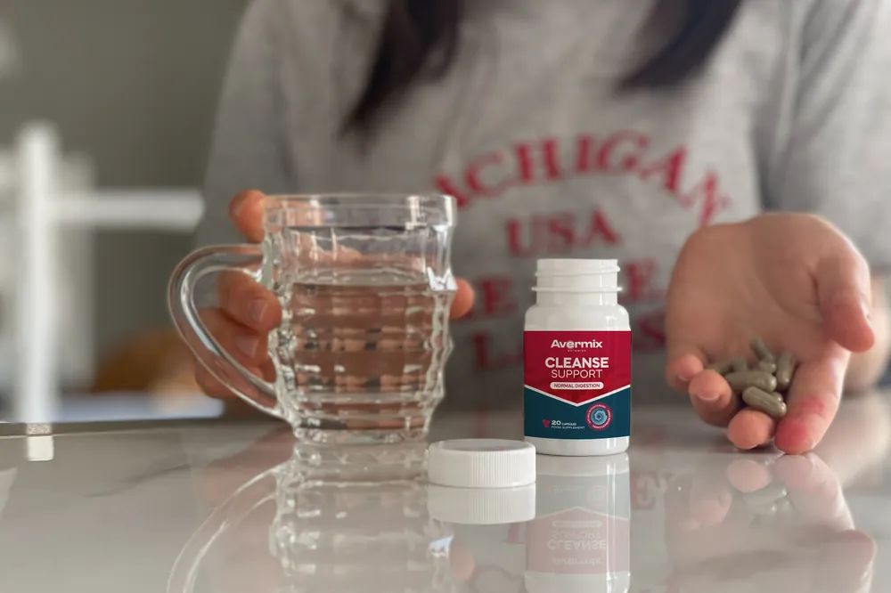 Donna a un tavolo con un bicchiere d'acqua, capsule Avermix nel palmo e il flacone accanto